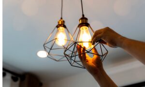 Hands adjusting a filament light bulb inside a geometric wire fixture, with another similar lit fixture nearby in a modern interior setting.