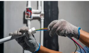 A person wearing gloves connects colored electrical wires to a white conduit.