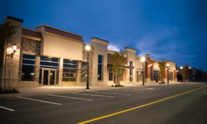Row of modern, empty storefronts with large glass windows, lit by streetlights at dusk, with an empty parking lot in the foreground.