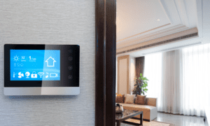 Smart home control panel on a wall displaying weather, date, and home settings, with a modern living room visible in the background.