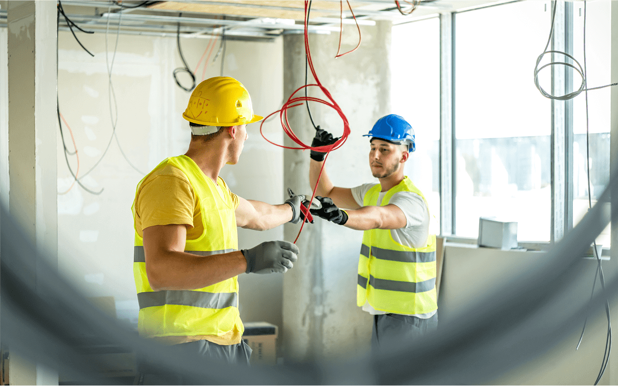 Two construction workers wearing helmets and safety vests are installing electrical wires in a building under construction.