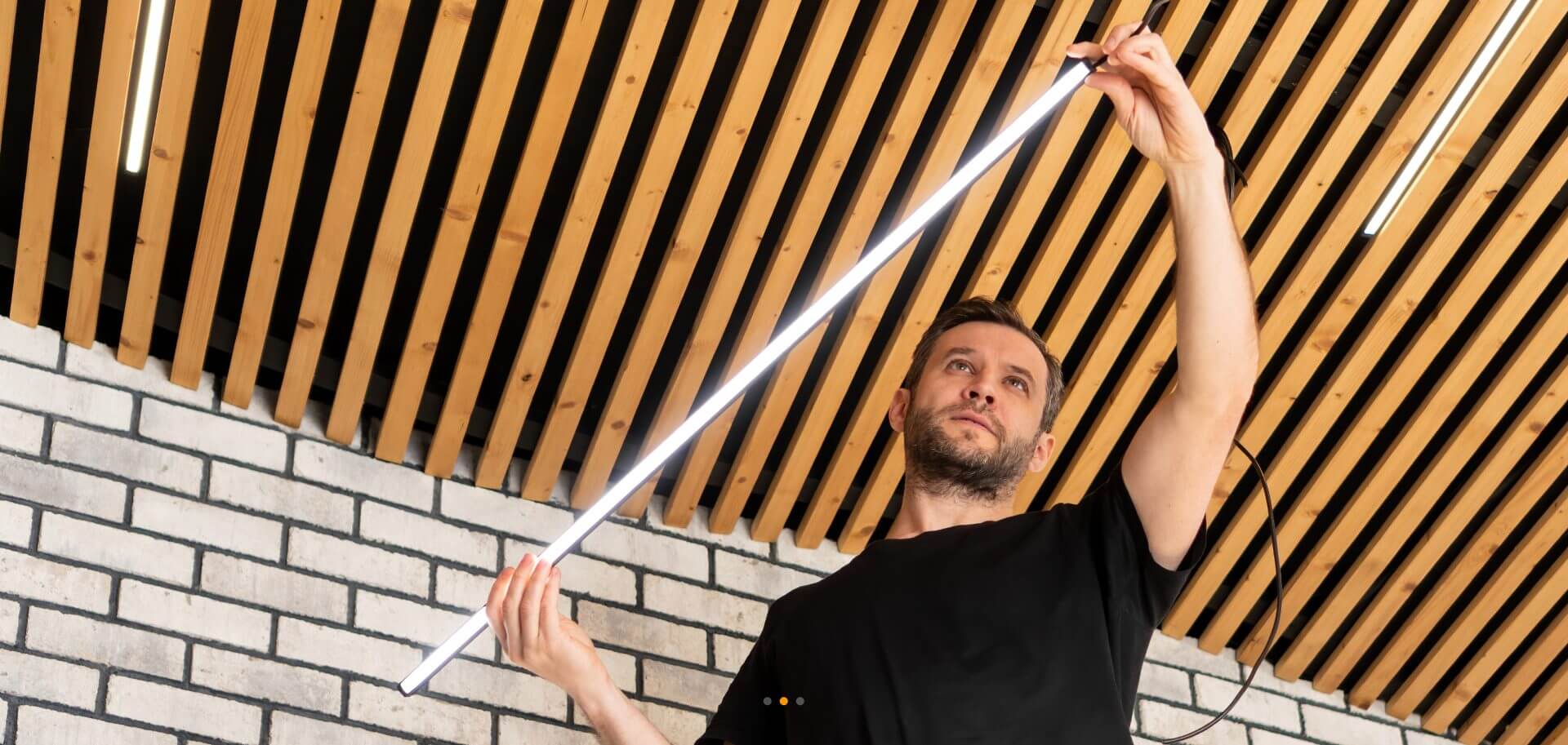 Man in a black shirt installing an LED light fixture on a wooden slatted ceiling in front of a white brick wall.
