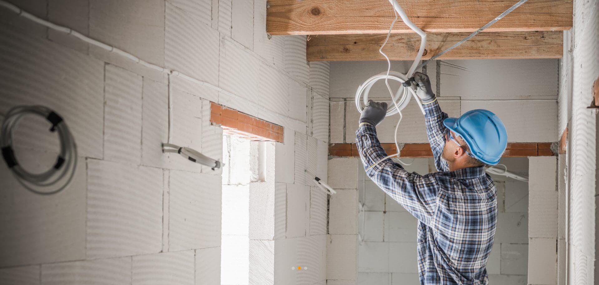 Electrician wearing a blue helmet and plaid shirt installing wiring on a ceiling in a building under construction.